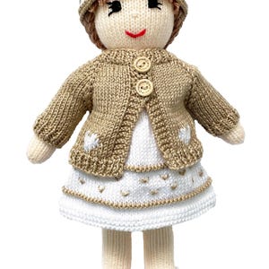 May include: A handmade knitted doll wearing a tan cardigan with wooden buttons, a white dress, and a matching hat. The doll has a smiling face, black embroidered eyes, and white shoes with gold accents. The doll is approximately 10 inches tall.