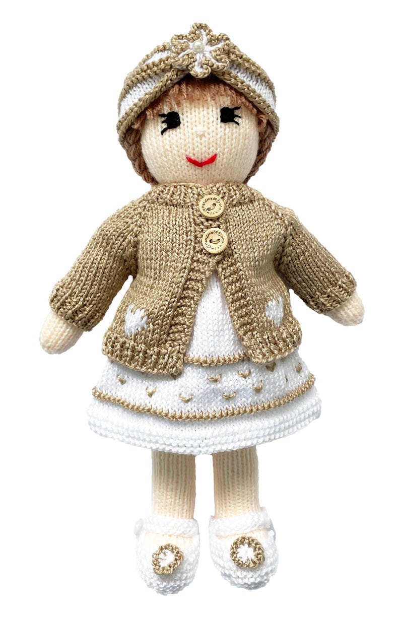 May include: A handmade knitted doll wearing a tan cardigan with wooden buttons, a white dress, and a matching hat. The doll has a smiling face, black embroidered eyes, and white shoes with gold accents. The doll is approximately 10 inches tall.