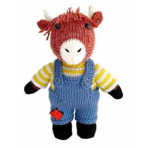 Pode incluir: Um peluche tricotado que representa uma vaca das Terras Altas com cabeça cor de ferrugem, focinho branco e pequenos chifres. Veste uma camisola de manga comprida às riscas amarelas e brancas sob jardineiras de malha azul com dois botões.