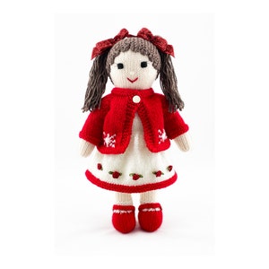 May include: A knitted doll with brown hair and red bows. The doll is dressed in a red cardigan with white snowflake patterns, a white dress with red rose embroidery, and red shoes. The doll is approximately 30 cm tall.