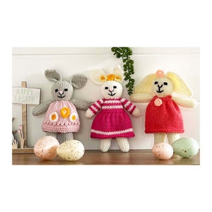 May include: Three knitted bunny rabbits wearing dresses. The bunnies are gray, white, and yellow. The dresses are pink, red, and pink with white stripes. The bunnies are sitting on a wooden surface with Easter eggs.