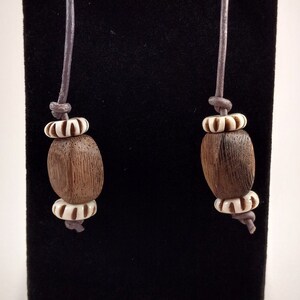 May include: A pair of brown wooden bead earrings with white accents hanging from gray leather cords on a black background.