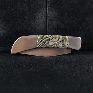 Könnte beinhalten: Ein kleines, zusammenklappbares Taschenmesser mit einer Klinge und einem Griff aus Edelstahl. Der Griff weist ein einzigartiges Muster aus grünen und weißen Wirbeln auf. Das Messer ist geschlossen, die Klinge ist im Griff verstaut. Das Messer liegt auf einer schwarzen Oberfläche.