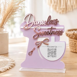 May include: A lavender-colored stand mixer-shaped sign with the words "Daniela's Sweetness" in rose gold script. A small whisk and bow are also included. The sign has a clear base and includes a "Follow Us" section with a QR code.