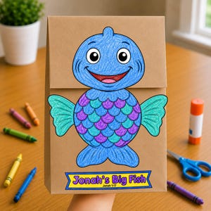 May include: A brown paper bag craft featuring a hand-colored blue fish with purple and teal scales. The fish has a smiling face and large eyes. Below the fish is a yellow banner with a blue border containing the text Jonah's Big Fish Jonah 1:17.