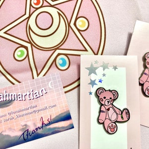 K&ouml;nnte beinhalten: Zwei rosa Teddyb&auml;r-Pins mit verbundenen Armen und Beinen. Die Pins befinden sich auf einem wei&szlig;en Hintergrund mit einer rosa und wei&szlig;en Karte mit Sternen und dem Text "Lena Martian" sowie Kontaktinformationen.