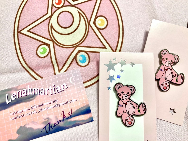 K&ouml;nnte beinhalten: Zwei rosa Teddyb&auml;r-Pins mit verbundenen Armen und Beinen. Die Pins befinden sich auf einem wei&szlig;en Hintergrund mit einer rosa und wei&szlig;en Karte mit Sternen und dem Text "Lena Martian" sowie Kontaktinformationen.