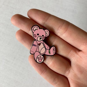 K&ouml;nnte beinhalten: Eine rosa Teddyb&auml;r-Anstecknadel mit einem Herz auf der Brust und einem Verband am Arm. Der B&auml;r hat schwarze Augen und eine schwarze Nase. Die Anstecknadel ist aus Metall und hat eine gl&auml;nzende Oberfl&auml;che.