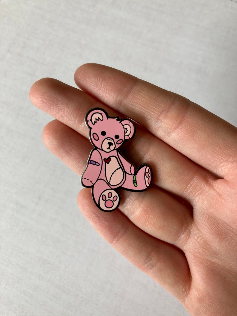 K&ouml;nnte beinhalten: Eine rosa Teddyb&auml;r-Anstecknadel mit einem Herz auf der Brust und einem Verband am Arm. Der B&auml;r hat schwarze Augen und eine schwarze Nase. Die Anstecknadel ist aus Metall und hat eine gl&auml;nzende Oberfl&auml;che.