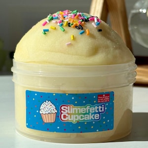 May include: A jar of yellow slime topped with colourful sprinkles. The jar features a blue label with a cupcake graphic and the words "Slimefetti Cupcake". A warning label states "This product is not edible. Do not eat it."