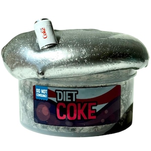 May include: A clear plastic container filled with small, silver-colored pieces, topped with a large, silver-colored blob. A miniature Diet Coke can is embedded in the blob. The container has a label that reads "DO NOT CONSUME" and "DIET COKE."