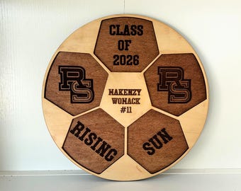 May include: A wooden soccer ball-shaped plaque with laser-cut details. The design includes the text "CLASS OF 2026", "HAKENZY WOMACK #11", "RISING", and "SUN". The plaque features a light wood base with darker wood accents.