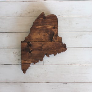 May include: A wooden cutout of the state of Maine, stained in a dark brown color. The cutout is on a white wood background.