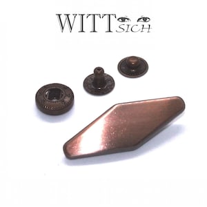 Könnte beinhalten: Kupferfarbene Metallbeschläge, darunter Druckknöpfe und ein rautenförmiger Verschluss, sind auf weißem Hintergrund angeordnet. Der Schriftzug "WITT SICH" ist zu sehen. Die Beschläge haben eine Vintage-Optik.