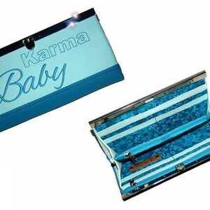Op de afbeelding: Een lichtblauwe clutch portemonnee met de woorden "Karma Baby" geborduurd op de voorkant. De portemonnee heeft een zilveren sluiting en een donkerblauwe rand. De binnenkant heeft een gestreept patroon met een bloemmotief en een vak met ritssluiting.