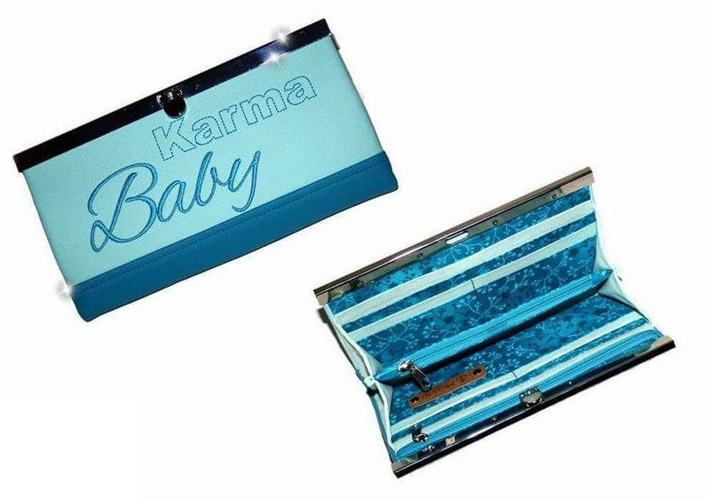 Op de afbeelding: Een lichtblauwe clutch portemonnee met de woorden "Karma Baby" geborduurd op de voorkant. De portemonnee heeft een zilveren sluiting en een donkerblauwe rand. De binnenkant heeft een gestreept patroon met een bloemmotief en een vak met ritssluiting.