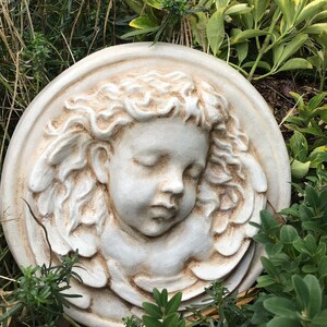 May include: A white ceramic plaque with a sculpted cherub face with closed eyes and a halo of hair. The plaque is surrounded by a raised border and has a distressed white finish.