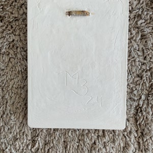 May include: White rectangular plaque with a gold metal hanging hook. The plaque has a handwritten inscription that reads "M3 24".