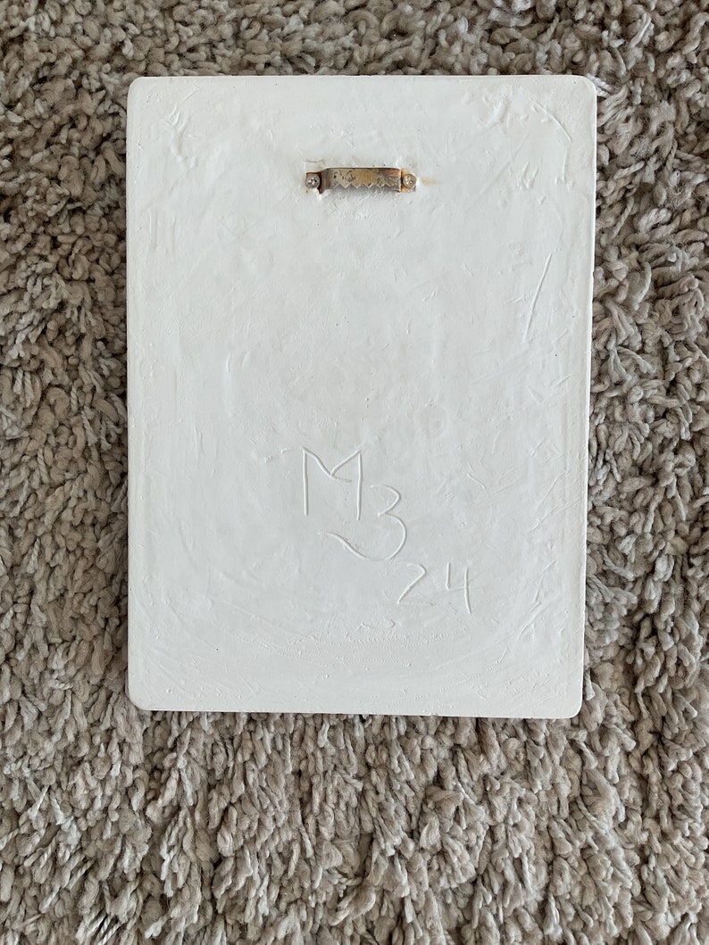 May include: White rectangular plaque with a gold metal hanging hook. The plaque has a handwritten inscription that reads "M3 24".
