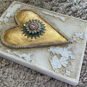 May include: A gold heart-shaped sculpture with a jeweled center, surrounded by a row of rhinestones, on a white background with a distressed finish.