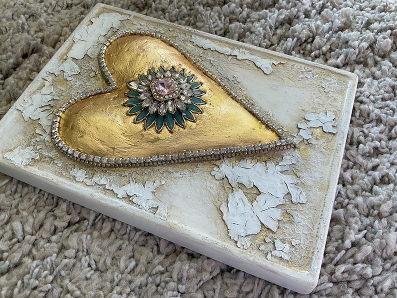 May include: A gold heart-shaped sculpture with a jeweled center, surrounded by a row of rhinestones, on a white background with a distressed finish.