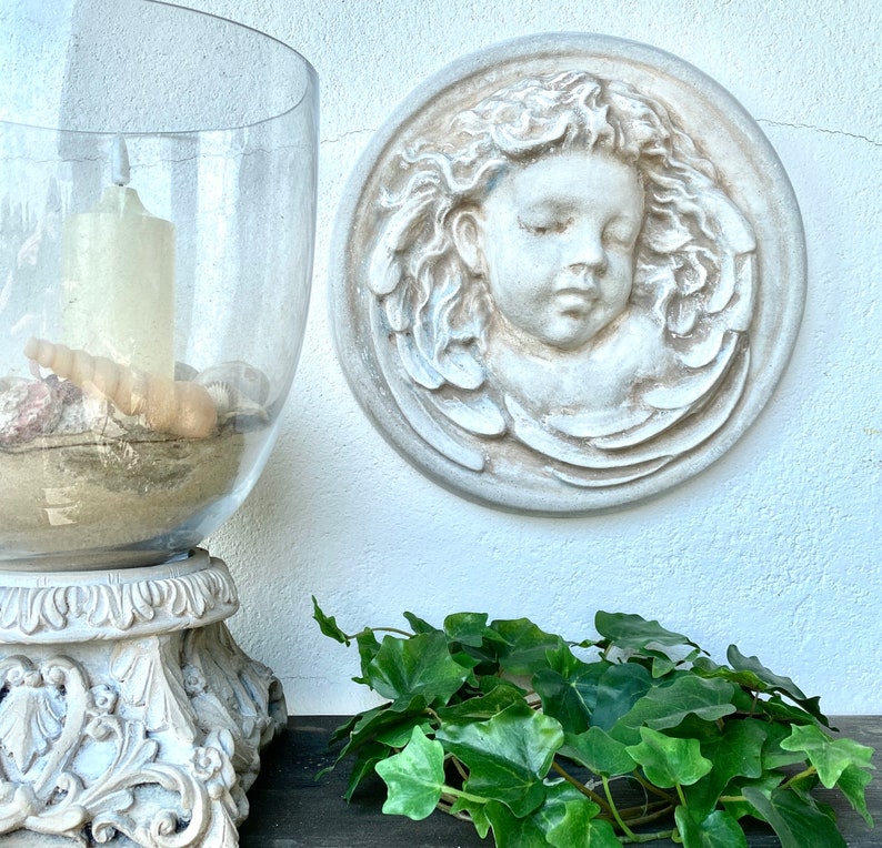 May include: A white plaster wall decor featuring a cherub with closed eyes, surrounded by a decorative floral design. The decor is mounted on a white wall, with a glass candle holder and green ivy plants in the foreground.