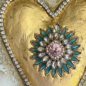 May include: A gold heart-shaped ornament with a large, jeweled flower design in the center. The flower has a pink center surrounded by clear and green rhinestones.