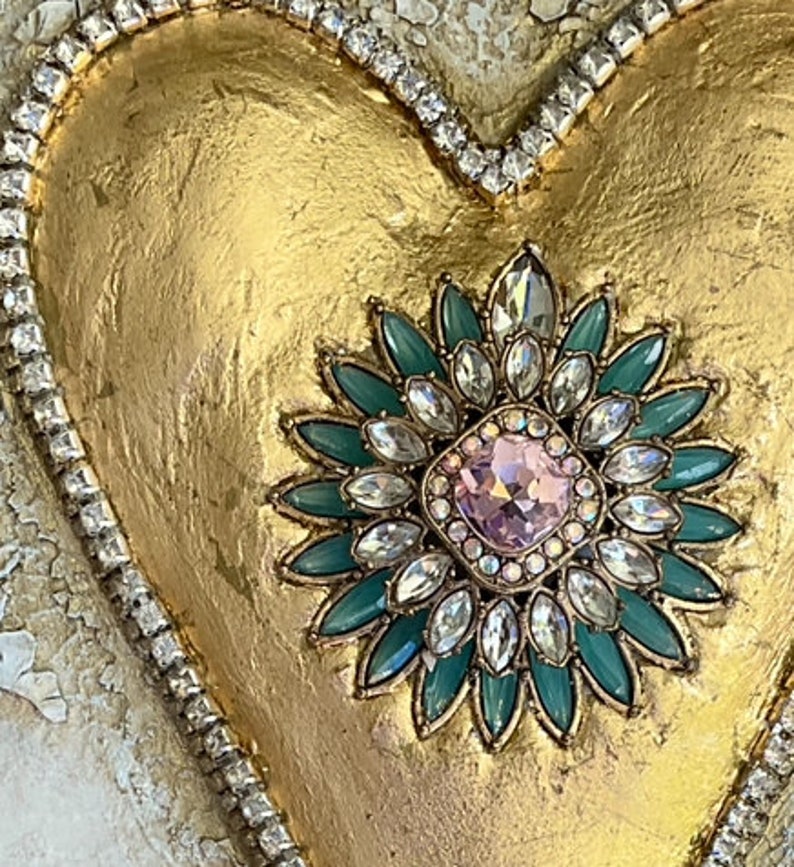 May include: A gold heart-shaped ornament with a large, jeweled flower design in the center. The flower has a pink center surrounded by clear and green rhinestones.