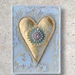 May include: A gold heart with a jeweled center on a white textured background. The heart is decorated with rhinestones and a green and pink flower with a large pink rhinestone in the center.
