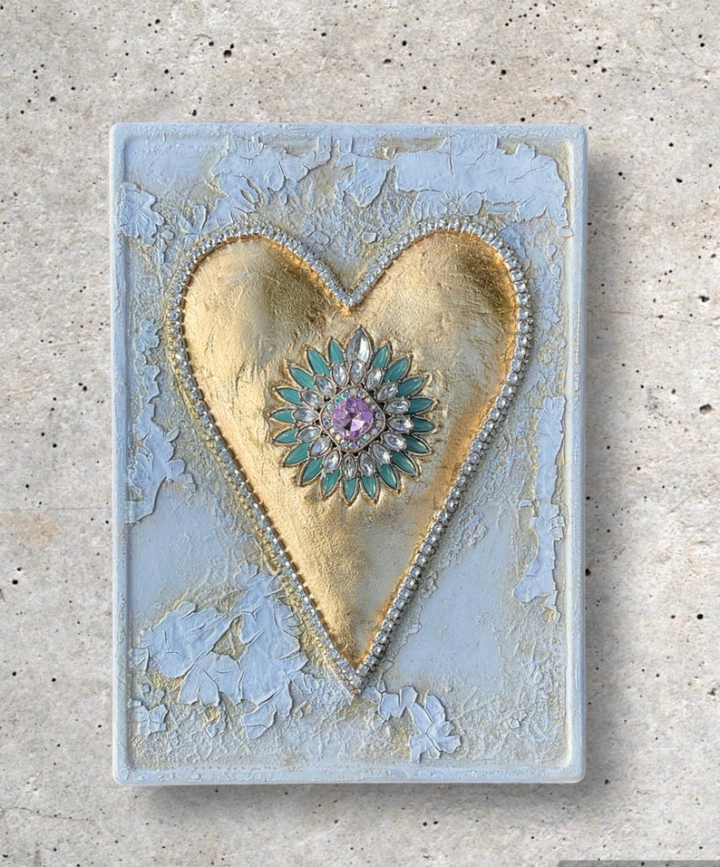 May include: A gold heart with a jeweled center on a white textured background. The heart is decorated with rhinestones and a green and pink flower with a large pink rhinestone in the center.