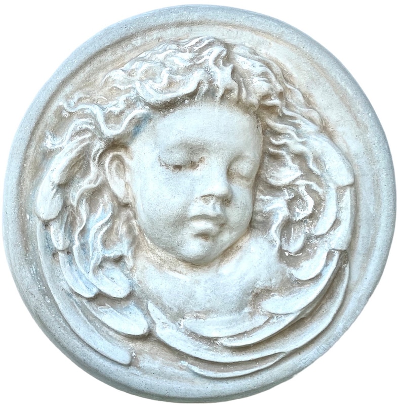 May include: A white ceramic plaque with a bas-relief sculpture of a cherub's face with closed eyes and wings.