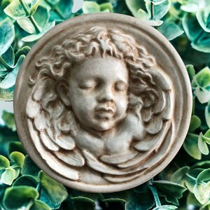 May include: A white ceramic sculpture of an angel with closed eyes and wings. The sculpture is in a circular frame and has a distressed finish.