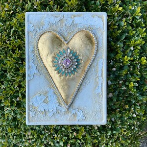 May include: A white square canvas with a gold heart shape with a green and pink jeweled center. The heart is surrounded by a row of small white rhinestones.