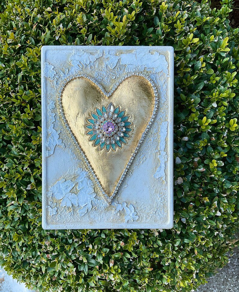 May include: A white square canvas with a gold heart shape with a green and pink jeweled center. The heart is surrounded by a row of small white rhinestones.