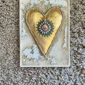 May include: A gold heart-shaped plaque with a jeweled embellishment in the center. The plaque has a distressed white background and is framed with a beaded border.