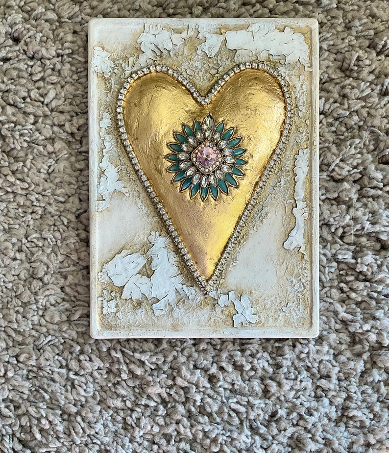 May include: A gold heart-shaped plaque with a jeweled embellishment in the center. The plaque has a distressed white background and is framed with a beaded border.
