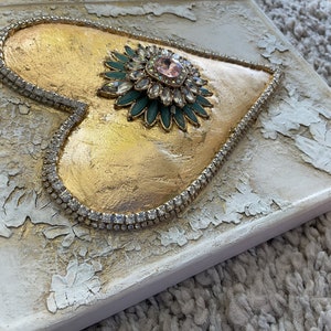 May include: A gold heart-shaped canvas with a large, sparkly, green and pink jeweled embellishment in the center. The heart is surrounded by a row of clear rhinestones.