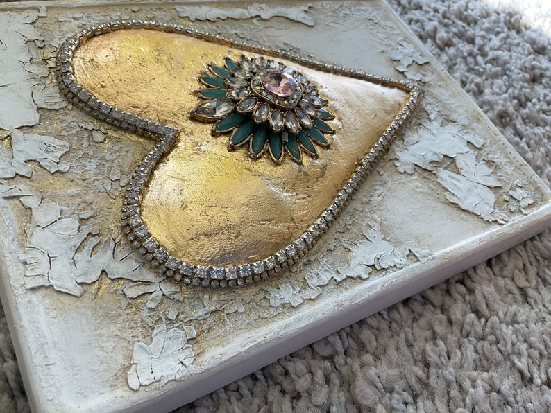 May include: A gold heart-shaped canvas with a large, sparkly, green and pink jeweled embellishment in the center. The heart is surrounded by a row of clear rhinestones.