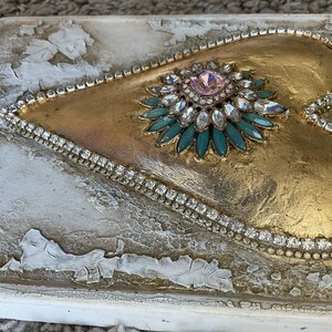 May include: A gold heart-shaped canvas with a large jeweled embellishment in the center. The embellishment is a flower-like design with a pink center stone surrounded by clear and teal rhinestones.