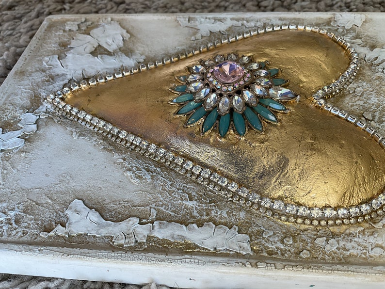 May include: A gold heart-shaped canvas with a large jeweled embellishment in the center. The embellishment is a flower-like design with a pink center stone surrounded by clear and teal rhinestones.