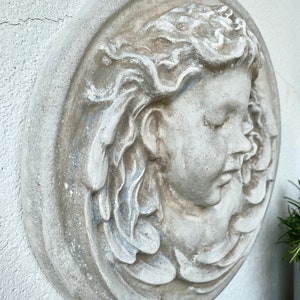 May include: A white plaster wall sculpture of a woman's face with flowing hair. The sculpture is in a round frame and has a distressed white finish.