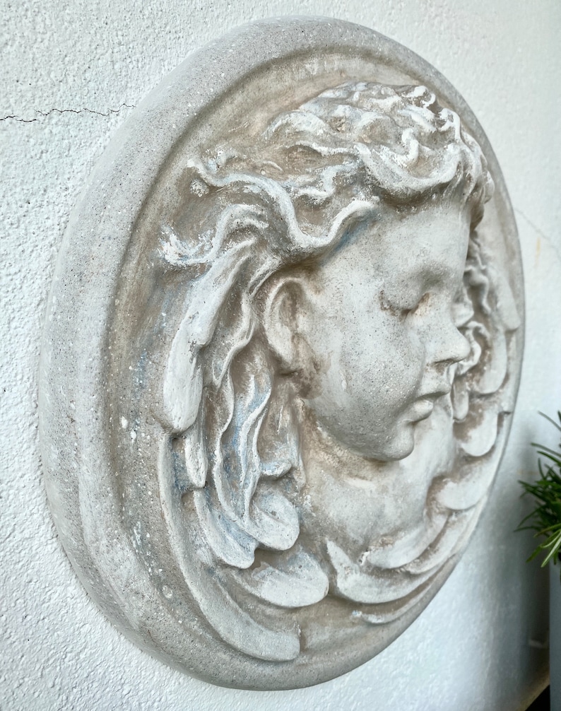 May include: A white plaster wall sculpture of a woman's face with flowing hair. The sculpture is in a round frame and has a distressed white finish.