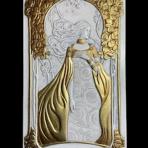 May include: A rectangular, Art Nouveau-style relief sculpture. The artwork features a woman with long flowing hair and a gold-colored dress. The relief is primarily white with gold accents, including floral and decorative border elements. The background has a swirling pattern.