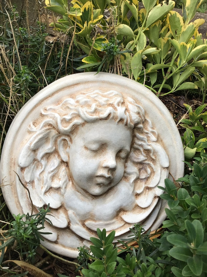 May include: A white ceramic plaque with a sculpted cherub face with closed eyes and a halo of hair. The plaque is surrounded by a raised border and has a distressed white finish.