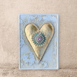May include: A gold heart with a green and pink jeweled center, on a white textured background.