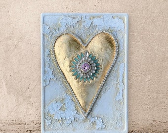 May include: A gold heart with a green and pink jeweled center, on a white textured background.