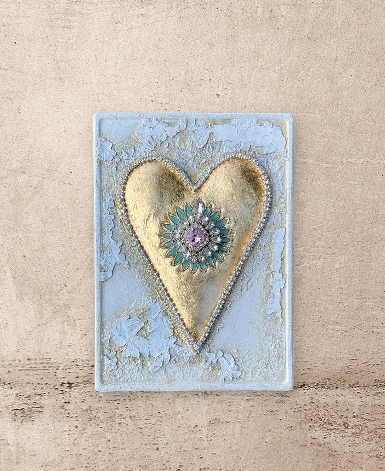 May include: A gold heart with a green and pink jeweled center, on a white textured background.