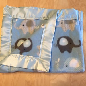 May include: A light blue baby blanket with a satin border. The blanket features a repeating pattern of gray and white elephants on a light blue background.