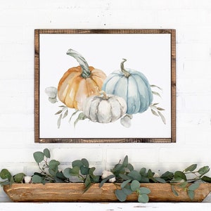 May include: A framed watercolor print featuring three pumpkins in shades of orange, blue, and white, with green foliage. The artwork is displayed above a white mantel with a wooden trough filled with greenery.