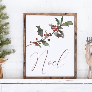 May include: A framed sign with the word "Noel" in elegant script, featuring a watercolor holly branch with red berries. Two small artificial Christmas trees and a wooden reindeer are on a white mantel.